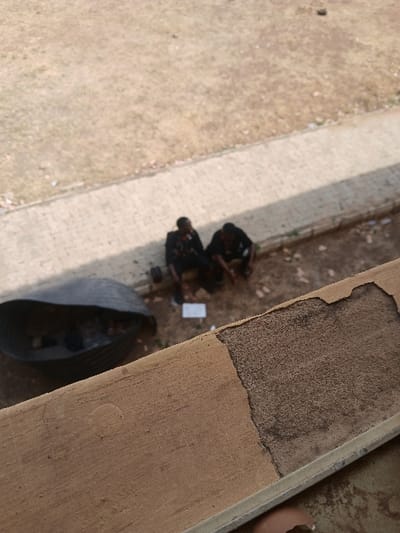 The image presents an elevated perspective of an outdoor scene in Ugi Majowel, Nigeria. In the foreground, a light brown concrete architectural element, possibly a ledge or wall, shows an irregular patch of its surface coating absent, revealing a darker, granular texture beneath.
Below this element, in the midground, two adult individuals are seated on dry, brownish ground. The individual on the left is dressed in a dark jacket over a lighter shirt, displaying indistinct insignia or patterns on the chest. The individual on the right wears dark clothing. Both individuals are looking downwards, appearing to focus on a light-colored, rectangular object (possibly paper or a small box) situated on the ground directly between them. To the left of these individuals, a large, dark, trough-shaped corrugated object, resembling a cut section of a pipe, rests on the ground and contains various unidentifiable items.
A paved pathway, constructed from light-colored pavers, runs diagonally behind the seated individuals. Beyond this pathway, the background consists of a broad expanse of dry, yellowish-brown ground with sparse vegetation. The scene is illuminated by daylight, and the midground elements, including the individuals, appear slightly out of focus.