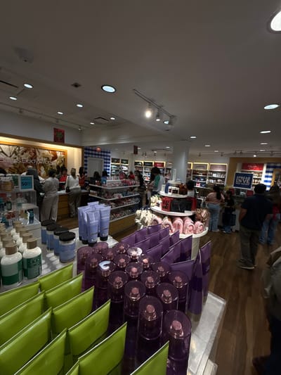 Inside a brightly lit Bath & Body Works store, customers browse merchandise. Shelves display candles, lotions, and other fragrant items. A large display features purple and green products, creating a visually appealing arrangement. People can be seen shopping in the aisles, adding to the busy atmosphere of the retail space.