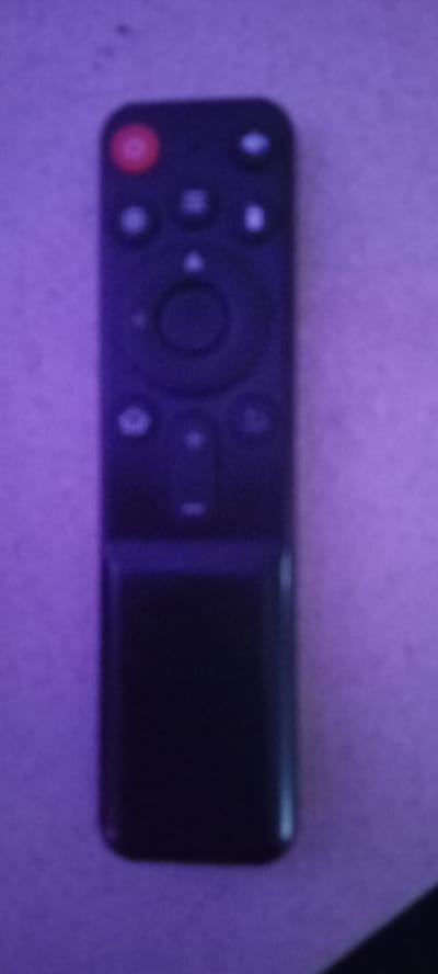 The image shows a black remote control with several buttons. The top button is red and appears to be a power button. Below that are several circular buttons, some with symbols. The bottom portion of the remote is a solid black rectangular area. The remote is placed on a textured surface that appears to be a light-colored fabric or wall, illuminated by purple light. The image quality is low, with significant blurriness. There is no information about the city of Perekip, Ukraine, in this image.