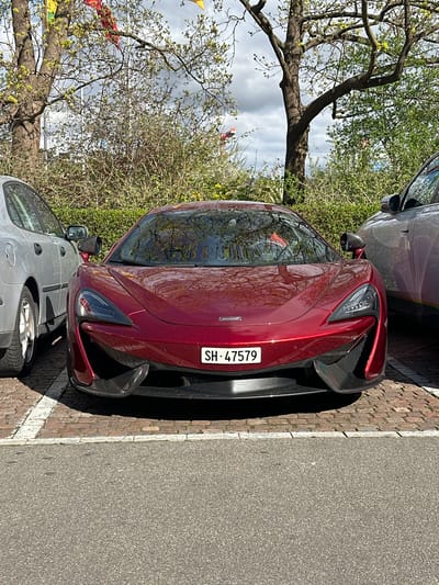 The image shows a maroon McLaren Artura parked on a brick-paved section of a street in Schaffhausen, Switzerland. The car has a license plate that reads "SH-47579." It is parked next to a gray vehicle, likely a car, and near a sidewalk marked with a white line. A tree with budding leaves is visible in the background, along with other greenery. The sky is partly cloudy. The pavement adjacent to the brickwork appears to be asphalt.