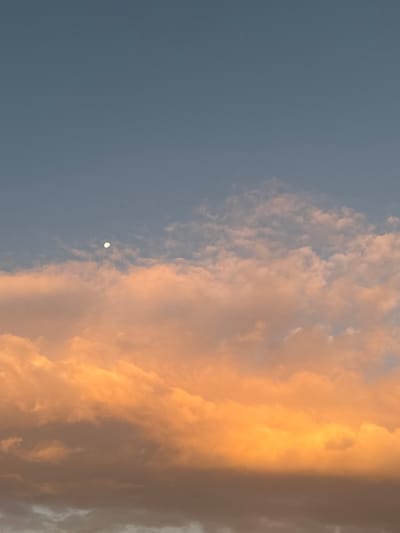 A bright moon punctuates the pale blue sky to the left. The right side is filled with thick clouds, painted with shades of orange and gold by a setting sun. The contrast between the cool, dark blue of the sky and the warm, luminous clouds creates a dramatic scene. Wisps of white cloud peek through the blue expanse.