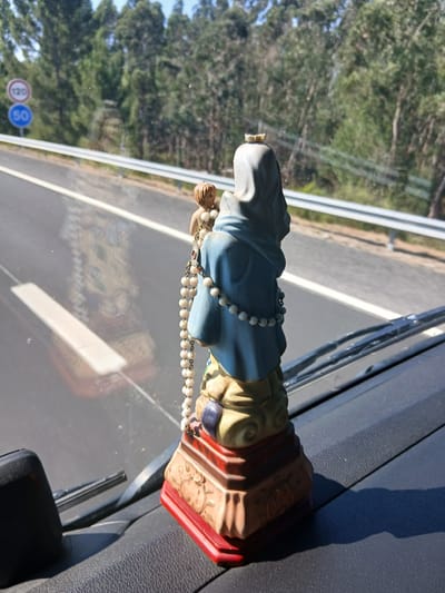 A statue of the Virgin Mary holding a child stands on the dashboard of a vehicle, positioned on the right side of the frame. The statue is adorned with a string of white beads, resembling a rosary, draped around its neck and shoulders. A small golden crown rests atop the Virgin Mary's head. The statue is mounted on a multi-colored base, predominantly red and beige, with decorative elements visible.

Through the windshield, a highway is visible, bordered by a white guardrail. On the left side of the highway, two circular traffic signs are displayed: one with "120" in black on a white background, and another below it with "50" in white on a blue background. Beyond the highway, a dense forest of tall, green trees forms the backdrop. The sky appears clear and bright. A reflection of the statue and possibly the interior of the vehicle is visible on the windshield.

The image was captured from within the vehicle, looking outwards through the windshield. The dashboard and windshield wipers are partially visible in the foreground. The overall scene suggests a journey or travel, likely on a highway. The location of Rio Maior, Portugal is provided as context for the scene.