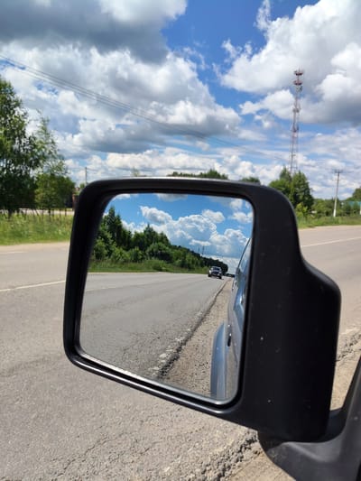 The image is a photograph taken from inside a vehicle, looking out through a side mirror. The mirror reflects a road with trees and a blue sky with white clouds. A dark-colored car is visible on the road in the reflection. The main image shows the side of the vehicle and the road with power lines and a cell tower in the background. The setting appears to be a rural or suburban area. The location is specified as Khodaevo, Russia.