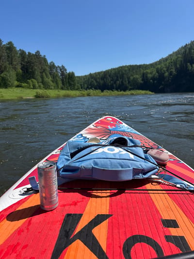 This is a first-person perspective shot from a paddleboard on the Mana River in Manskii, Russia, under a clear blue sky. The paddleboard has a colorful, fish-themed design. In the foreground, resting on the paddleboard, are a blue backpack, a silver can of what appears to be sparkling water, and a small, beige portable speaker. The river is calm with visible ripples, and the banks are lined with dense, green coniferous forests. The water reflects the sunlight, creating sparkling highlights.