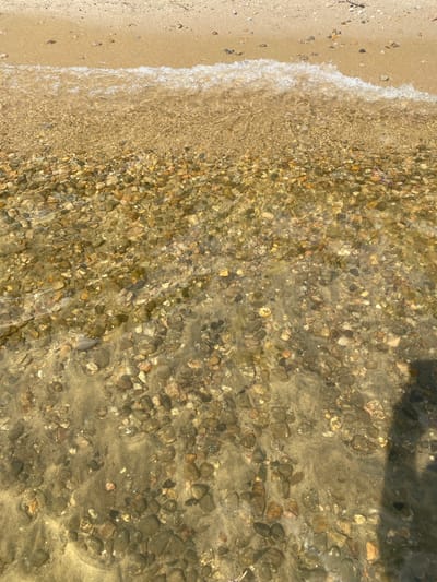 The image shows a shallow body of clear water lapping against a sandy and pebbly shore. The water is transparent, revealing a bed of smooth, multi-colored stones and sand. A thin wave crests at the top of the frame, composed of white foam. The shore is composed of tan sand with scattered small pebbles and some organic debris. The overall scene suggests a natural waterfront environment, possibly a beach or lake shore. No specific landmarks or details are visible to pinpoint a location within Duhamel, Canada.