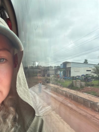 The image is a selfie taken from inside a vehicle, looking out through a window. The left side of the frame shows the face of a person wearing a hooded garment. The rest of the image captures an outdoor scene with a building, construction materials, and a muddy road. The building appears to be a commercial or industrial structure with a light-colored facade and a metal roof. In front of the building, various pipes and containers are stored, suggesting a construction or manufacturing site. The road is unpaved and appears wet and muddy. The sky is overcast, with visible power lines stretching across the frame. The general impression is a utilitarian or industrial area under a cloudy sky. Based on the context provided that the location is the city of Phao, Laos, this scene depicts an outdoor industrial or construction area in that region.