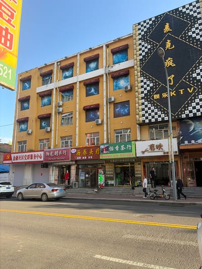 The image shows a street scene in Aihui, China. On the left is a tall, yellow building with multiple floors. Several windows on the upper floors are decorated with images of galaxies or nebulae. Red awnings are above many of these windows. Below these windows, on the ground floor, are various shops with signs in Chinese. One sign reads "金融社区党群服务中心" (Financial Community Party Mass Service Center), another "阳光刺衣行" (Sunshine Clothing Store), and another "汤养头疗" (Soup Health Head Therapy).

To the right of the yellow building is a taller, dark building with a large, ornate diamond-shaped design composed of black and white squares and gold accents. Large yellow Chinese characters are visible on this building, along with the letters "KTV".

In front of the buildings, a silver sedan is parked on the street. The street is paved and has double yellow lines. Sidewalks are in front of the shops. Several people are visible on the sidewalk. One person is standing in the doorway of a shop, and others are walking along the sidewalk. A scooter is parked on the sidewalk. The sky is clear and blue. A yellow sign with red Chinese characters is on the far left edge of the image.