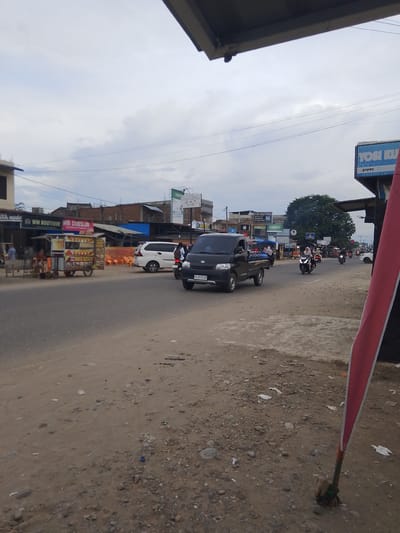 The image depicts a street scene in Gampongpaya, Indonesia. A black pickup truck is driving on the right side of the road. To the left of the truck, a white minivan is parked. In the background, there are several buildings and a large tree. The sky is overcast. A red and white flag is visible in the foreground. The road appears to be paved, with a dirt shoulder on the right side. There are several people on motorcycles and a food cart on the left side of the road. Signs for "WM BOUTIQUE" and "YOSI KU" are visible on the buildings. The overall impression is a busy street in a small town or city.