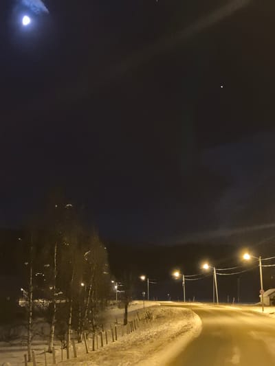 The image displays a nighttime winter scene in Leiknes, Norway. A two-lane asphalt road, partially covered in snow and ice, curves gently to the right from the foreground. The road and its immediate surroundings are illuminated by a series of amber-toned streetlights mounted on utility poles spaced along the right side. On the left, a snow-covered embankment is lined with a row of slender, deciduous trees, mostly bare, behind a low wooden fence. The ground throughout the visible area is covered in snow. The dark night sky occupies the upper portion of the frame, featuring a bright, crescent moon in the upper left quadrant, partially obscured by light clouds, and a singular, distinct bright point of light, possibly a star or planet, in the upper right. On the far right side of the road, a small, snow-dusted structure is visible, adjacent to a circular red and white speed limit sign displaying "60". No people, animals, vehicles, or observable actions or interactions are present in the scene.