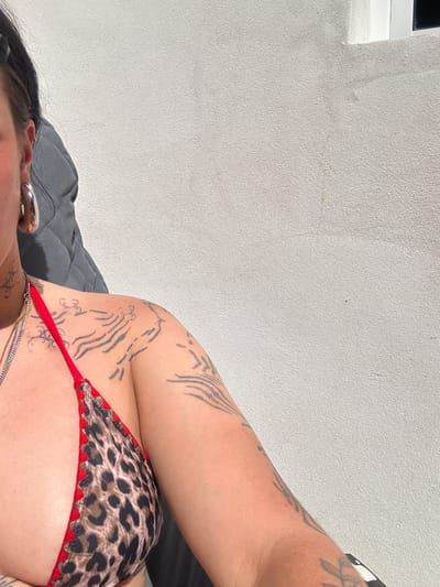 The image shows a person lying down, possibly outdoors in La Couarde-sur-Mer, France, given the context. The person has dark hair and visible tattoos on their arms and chest. They are wearing a leopard print bikini top with red trim. A silver ear gauge is visible in their left ear. The background features a light-colored textured wall and a gray fabric surface, possibly a cushion or bed.