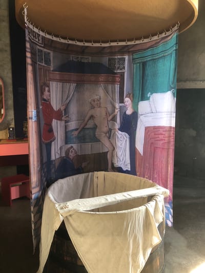 The image shows a tapestry depicting a historical scene, displayed in what appears to be a museum or historical site. The tapestry portrays a medieval setting, with figures in period clothing; a woman stands beside a reclining or deceased male figure, attended by other people. The scene includes a bed with green, white, and red coverings. The tapestry is suspended by a wooden frame with hooks. A section of folded white cloth or canvas partially obscures the right side of the tapestry. The display is in a dimly lit indoor environment, likely within a historical structure in Veytaux, Switzerland. The city name is not visible within the tapestry; the location name comes from the image caption. A small part of a pink colored object, likely a piece of furniture, is visible in the lower right.