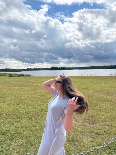 In Мазалаўскі сельскі Савет, Belarus, on a cloudy day, a young woman with long brown hair and sunglasses stands in a grassy field. She is wearing a grey dress with white stripes on the side. Her right hand is raised and touching her hair, which is blowing in the wind. In the background, there is a large body of water, likely a lake, with a treeline on the opposite shore. Several small figures are visible in the water and on the shore, suggesting a recreational area. A metal chain barrier is visible in the foreground, partially obscuring the bottom right corner of the image.