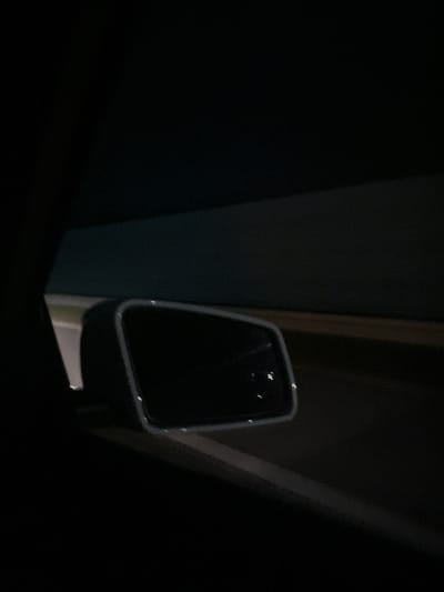 The image depicts a side-view mirror of a vehicle, possibly inside a garage or a dimly lit parking structure. The surroundings are predominantly dark, with vertical structural elements visible, likely supporting beams or walls. There are no identifiable people, subjects, actions, or interactions within the frame. The image lacks sufficient detail to determine a specific location in Djerman, Bulgaria, or any other location.