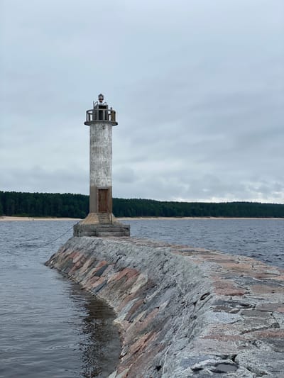 The image shows a concrete lighthouse situated on a stone pier extending into a body of water under an overcast sky. The lighthouse is cylindrical with a weathered, grey exterior and a metal railing structure at its top, which houses the lamp. A rusty metal door is visible on the lower part of the lighthouse, accessible by a short flight of stone steps. The pier is constructed from large, irregularly shaped stones of varying colors, including grey, pink, and reddish hues, and is lined with what appears to be white rope or cable. The water is dark and choppy, with ripples indicating a slight breeze. In the background, a dense forest of green trees lines a sandy beach. The overall scene suggests a remote coastal or lakeside location. The provided crop hints that the lighthouse may be located near Моторное, Russia.