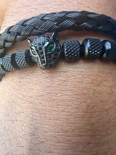 The image shows a close-up of a person's arm wearing a bracelet. The bracelet is made of black braided material and features several black metallic beads with a textured, knurled design. A prominent detail of the bracelet is a black panther's head charm, adorned with small black stones and green eyes. The background is the person's skin, showing texture and fine hairs. The lighting suggests it is daytime, and the skin tone and texture do not provide specific location cues. There is no visible text in the image. The focus is solely on the bracelet and the arm it is worn on, with no other subjects, activities, or setting details identifiable.