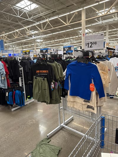 This image captures a retail clothing section within a large store, characterized by its high ceiling with exposed metal trusses and fluorescent strip lighting. The concrete floor and extensive shelving units suggest a big-box retailer or supermarket environment.

Numerous clothing racks display a variety of garments, predominantly children's or youth wear. In the upper-right portion, a rack holds blue long-sleeve shirts, one with a "Wonder Nation" tag, alongside light-colored shirts featuring the text "MARIO". Below these, another display showcases olive green outfits, including a black shirt with "MINECRAFT" branding and green shorts labeled "FORTNITE". Further down, racks are filled with dark-colored shirts and shorts, and folded stacks of blue, red, and black apparel. A loose green t-shirt lies on the concrete floor to the far right.

Price signs are visible hanging from the ceiling structure. One prominent sign reads "$12.98" and is partially labeled "Summer Months". Other signs indicate prices like "$5.23" (under "Posters"), "$5.88", and "$7", with additional numbers such as "10" and "15" seen on other displays. No people are visible in the scene, which depicts a static retail merchandising display. The ample indoor lighting suggests it is daytime. The setting is consistent with a commercial establishment in Worcester, United States.