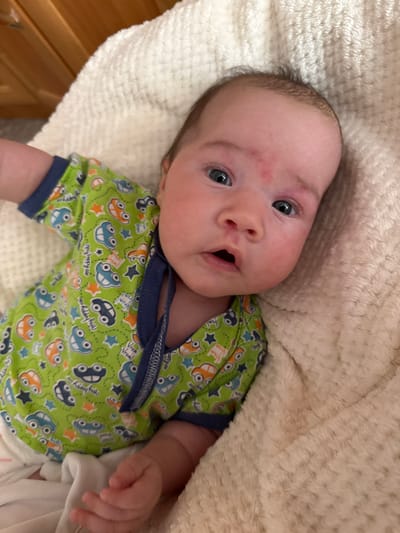 A newborn baby is lying on a cream-colored fluffy blanket, wearing a green onesie with a car print. The baby has a red mark on its forehead. The scene appears to be indoors, possibly in a nursery, given the presence of a wooden crib in the background. The lighting is warm and natural.