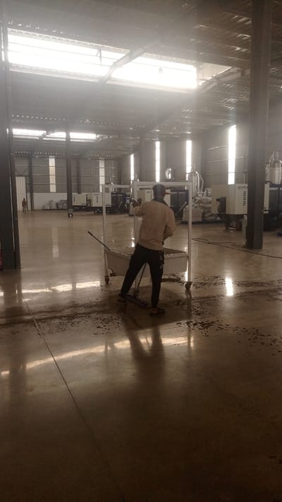 The image displays the interior of a large industrial facility located in Bwari, Nigeria. Natural light streams from multiple translucent skylights integrated into the corrugated metal roof structure. In the foreground, a male individual, wearing a light-colored long-sleeved shirt and dark trousers, is observed from behind, engaged in cleaning the floor. He is manipulating a blue-handled broom and a white, rectangular mobile cart with a trough, around which dark liquid stains and wet patches are visible on the smooth, reflective concrete floor. In the background, several light-colored industrial machines, some appearing to be marked with "HAITIAN" branding, are arranged along a wall, with bags of materials stacked nearby. The facility features dark structural support pillars and corrugated metal walls. A second, smaller figure is faintly visible in the far left background.