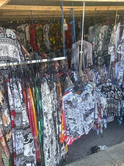 The image depicts a market stall in Bira, Indonesia, selling a variety of brightly colored and patterned clothing, predominantly short-sleeved shirts and possibly some dresses or skirts. The garments are hung from a wooden structure on multiple racks and suspended by blue cords. The patterns are varied, featuring tropical scenes, elephants, geometric designs, and abstract prints in black, white, red, green, yellow, and other vibrant hues. Some of the clothing appears to be made of light, breathable fabric, suggesting it is intended for a warm climate. In the lower right corner of the image, a calico cat is resting on a concrete surface, partially obscured by a low ledge. The ground beneath the stall is paved with concrete. The overall impression is of a bustling, colorful market setting characteristic of a tourist destination.