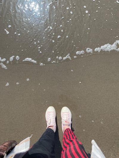 The scene depicts a pair of human legs and feet, viewed from an overhead perspective, standing on a dark, wet sandy beach in Ulcinj, Montenegro. The individual wears light pink sneakers, identified by visible Converse branding on the right shoe's medial side. The left leg is clothed in black pants, and the right leg is clothed in red and black vertically striped pants. A brown leather-textured object and a beige-and-white striped textile are partially visible on the left side of the frame. The sand transitions into ocean water in the upper third of the image, where a horizontal line of white sea foam and scattered bubbles indicates wave activity. Bright sunlight reflects off the water surface in the upper-left quadrant. A small, light-colored shell or pebble is visible on the sand near the foam line.