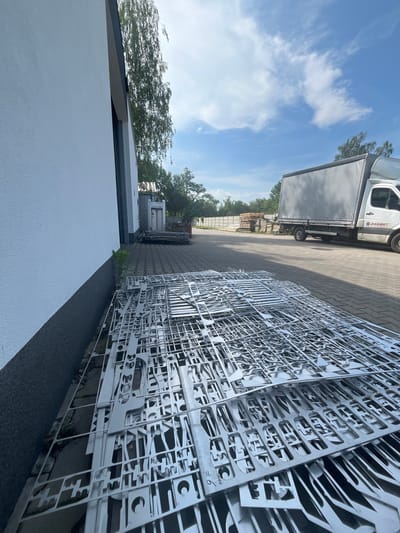 In Bogucin, Poland, an outdoor industrial setting is depicted. A long, white building runs along the left side of the frame, its textured wall dominating the foreground. In front of the building, a large pile of intricately cut, flat metal sheets lies on the ground, appearing to be remnants from a laser cutting or similar fabrication process. The metal pieces are arranged in a dense, overlapping layer. Beyond the metal sheets, a paved area stretches into the background. A white box truck with a grey tarp-covered cargo area is parked on the paved surface, its side bearing the logo "OSSMET". Further in the background, trees and a white fence are visible. The sky is blue with scattered white clouds. The scene suggests a materials processing or fabrication facility.