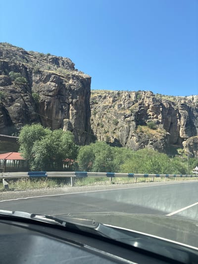 The image shows a road adjacent to a rocky mountain on a clear, sunny day. The road is paved and has a white line marking its center. A guardrail with blue and white striped posts separates the road from the grassy embankment. The mountain is steep and appears to be made of sedimentary rock, with visible layers and crevices. Patches of green vegetation are growing on the mountain slopes.

To the right of the road, a portion of a dark-colored vehicle is visible, including its windshield wiper and part of the dashboard. There is no visible text in the image. The lighting suggests it is daytime, likely morning or afternoon, given the clear blue sky and the bright sunlight illuminating the scene. The environment appears to be rural or mountainous, possibly a scenic route.