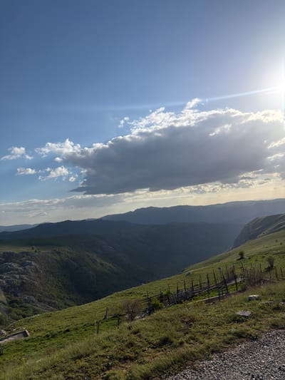 The image depicts a landscape near Plužine, Montenegro. The sky is blue with white clouds and a bright sun. A range of mountains extends into the distance. The foreground features a grassy hillside with a wooden fence and gravel.