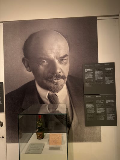 This media file is a photograph of a museum exhibit. The primary subject is a large black and white portrait of Vladimir Lenin, with his face angled and his gaze directed to the left. To the right of the portrait is a glass display case containing a small, green glass bottle with a red cap, and several documents. Behind the bottle and documents, a small framed text is visible.
In the upper right corner of the image, there are three rectangular plaques with text, likely providing information about the exhibit. The text on the plaques is visible and appears to be in at least three languages: German, French, and English. These plaques detail Lenin's time in Bern and Zurich, Switzerland, in 1914-1916.
The scene is indoors, likely in a museum or exhibition space, with plain walls and lighting that illuminates the display. The overall impression is one of historical documentation and preservation. The bottle and documents in the display case suggest personal effects or artifacts related to Lenin's life during his exile.
