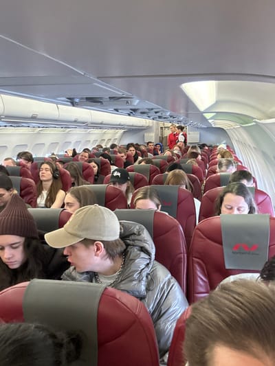 The image displays the interior cabin of a Nordwind Airlines commercial aircraft, with the indicated location being the city of Novogorelovo, Russia. The perspective is from a passenger seat, looking down the aisle towards the front and partially towards the rear. The cabin is brightly illuminated, with external daylight visible through windows on the left side of the aircraft.

Dozens of passengers occupy the dark red-upholstered seats, each featuring a gray headrest cover with the white "Nordwind Airlines" logo. Most passengers appear to be young adults, dressed in casual attire. In the immediate foreground, a male passenger wears a beige baseball cap, a shiny silver puffer jacket, and a silver chain around his neck; he is looking left with his mouth slightly open. To his left, a female passenger wears a dark brown beanie and dark clothing, looking downwards. Further back, a female passenger with long hair and white over-ear headphones has her eyes closed. Another male passenger in the mid-ground wears a black baseball cap with a white Stüssy logo. Passengers are mostly seated, some looking forward, others downwards, and some appear to be resting.

In the background, near the rear-right of the visible cabin, two female flight attendants are standing. They are dressed in red uniforms, with one blonde-haired attendant clearly visible, suggesting active duty or preparation. The overhead bins are closed and light grey, extending along both sides of the cabin.