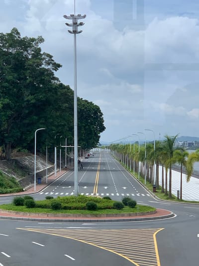 A wide asphalt road with yellow and white lane markings runs through a city. The road is lined with trees and streetlights on both sides. On the left side, a dense line of dark green trees borders a sloped embankment. On the right side, a line of palm trees separates the road from a pedestrian walkway with patterned paving stones. In the background, a body of water is visible, with buildings and hills on the far shore. The sky is cloudy, with large white clouds against a light blue background. The scene appears to be in a modern urban environment, possibly a coastal or riverside area, given the presence of water and tropical vegetation. This road is located in the Golden Triangle Special Economic Zone, Laos.
