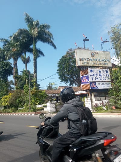 A person on a black motorcycle is driving on a road in front of the Sindanglaya Hotel-Restaurant, which is denoted by a large sign with small Indonesian flags on top. The sky is blue and sunny. Palm trees are visible on the left side of the image. A curb separates the road from the landscaping on the left. The sign for the hotel has Cafe S3 advertising below the hotel sign. The license plate is partially visible.