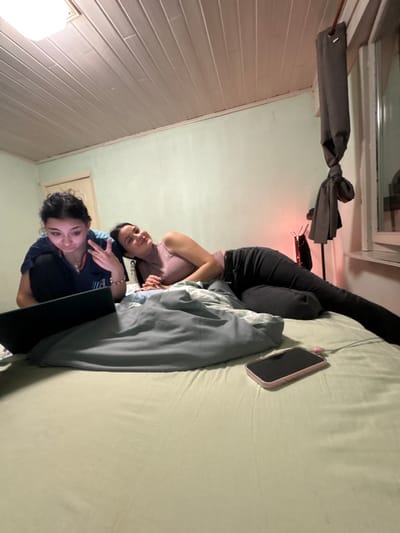Two female individuals are depicted inside a room, identified as located in Eeklo, Belgium. The scene appears to be a bedroom.

The first individual, on the left, has dark, curly hair and wears a dark blue top. She is positioned in a semi-upright posture on a bed, facing forward and looking at an open black laptop resting on the bed. Her right hand is raised, displaying a peace sign gesture. She wears a necklace and wrist bracelets.

The second individual, on the right, has dark, straight hair and wears a light purple long-sleeved top and dark trousers. She is lying on her left side on the bed, facing towards the first individual and the laptop, with a faint smile. Her legs are extended.

The bed is covered with a light green fitted sheet, and a light blue/grey duvet is partially between the individuals. A light pink smartphone, face down, with an attached white cable, is visible on the bed in the foreground.

The room features light green walls and a white paneled ceiling with a rectangular overhead light fixture. A wooden door frame is visible on the left background. On the right, a window is partially obscured by a dark grey curtain tied in a knot. A small, red-glowing light source on a stand is present behind the second individual. Lighting in the room is dim, supplemented by the laptop screen and the red light source.