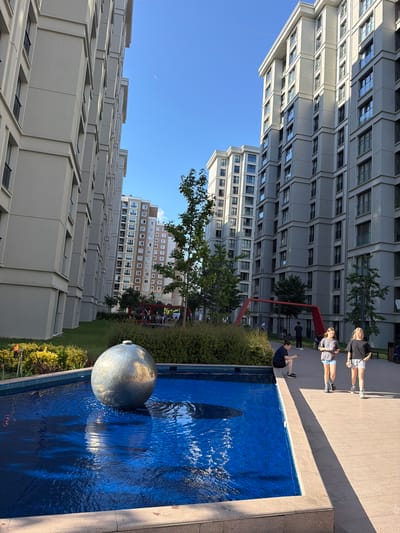 The image shows a contemporary urban courtyard in Zeytinburnu, Turkey, featuring a prominent spherical sculpture in a vibrant blue water fountain in the foreground. Tall, modern apartment buildings with repetitive window patterns flank the courtyard on either side, with one building on the left appearing closer and more in focus. The architecture is characterized by neutral tones of gray and beige. In the middle ground, a lush green lawn transitions to a densely planted area of shrubs and trees. A red abstract sculpture or play structure is visible in the background, with several people, including children, gathered around it. Towards the right side of the image, two teenage girls walk on a paved walkway, and a woman is seated near the fountain's edge. The sky is clear and blue, indicating fair weather. Sunlight casts distinct shadows across the pavement, highlighting the time of day.