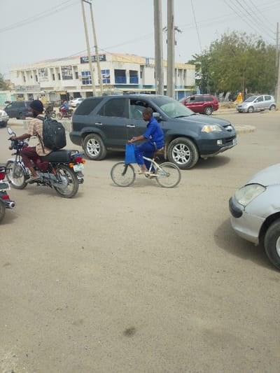 The image depicts a daytime street scene in Daurawa, Nigeria, characterized by vehicle and pedestrian traffic.
In the foreground, a light-colored motorcycle is positioned on the left, ridden by an individual wearing a light-brown patterned long-sleeved shirt, dark trousers, and a black backpack. The motorcycle bears a green Nigerian license plate (JIGAWA ASJ-64QJ). To the right, a young male, dressed in a blue short-sleeved shirt and blue trousers, is riding a small white bicycle with two wheels of seemingly equal size. He carries a blue plastic bag suspended from his left hand, near the handlebars.
Immediately to the right of the bicyclist, a dark-colored SUV, identifiable as an Acura MDX, is visible, oriented towards the right side of the frame. Its driver's side front window is partially lowered. The front section of a silver sedan is partially visible in the extreme lower right foreground.
The road surface is paved and appears to be made of light-grey asphalt or concrete, showing signs of wear and minor staining. Overhead, multiple utility poles with electrical wiring are present.
In the mid-ground and background, an off-white multi-story building with blue window frames and awnings is prominent. Signage on the building's facade includes "MEDIED SAHA VITAGE" and other partially obscured text, suggesting commercial activity. Several other vehicles, including a dark car, a red SUV, and a silver sedan, are visible further down the street. Green foliage from trees is visible on the right side of the background. The sky is bright and appears overcast or hazy.