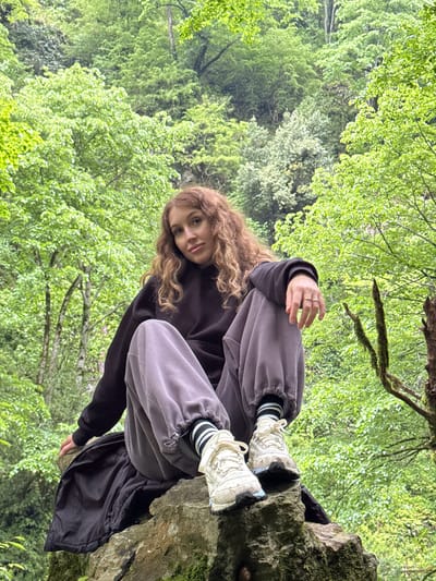 A young woman with curly brown hair is sitting cross-legged on a rocky outcrop in a forest setting. She is wearing gray sweatpants with black and white striped socks and white sneakers, and a dark-colored sweatshirt. She has rings on her fingers. The background consists of lush green foliage and trees, suggesting a natural environment. The image was taken in the city of Tsabal, Abkhazia.