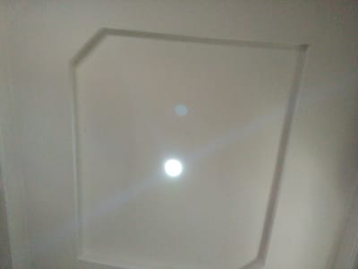 A ceiling in Rumuomasi, Nigeria, features a recessed, octagonal design with smooth, off-white surfaces. The perimeter of the recessed area is defined by a slightly raised molding. Centered within the recessed space are two circular lights. One light, located in the upper half of the recess, is dimly illuminated and appears as a soft, pale blue circle. The second light, positioned lower and more centrally, is brightly lit, emitting a strong white glow that casts subtle light rays onto the surrounding ceiling. The image is taken from a low angle, looking directly upwards.