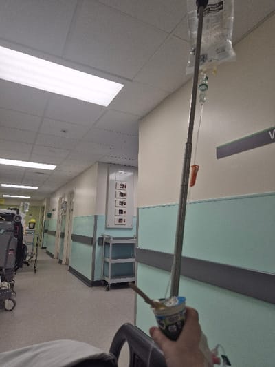 The scene depicts an interior view of a hospital corridor, likely within a medical facility in Hereford, United Kingdom. In the immediate foreground, a person's light-skinned hand, positioned at the bottom right, holds a white yogurt cup containing a spoon. An IV line is visible, trailing from near the hand. An adjustable metallic IV pole rises vertically from the lower right, supporting a clear IV fluid bag suspended from its top, with associated tubing and an orange clamp visible.
The corridor features off-white upper walls and a lower section painted light green, separated by a dark gray horizontal bumper rail. The ceiling consists of white acoustic tiles with recessed rectangular fluorescent light fixtures providing illumination. Multiple closed doors are visible along both sides of the hallway, receding into the distance. Medical equipment observed includes a three-tiered metallic utility cart against the right wall, a wall-mounted panel with eight white rectangular indicators further along the right wall, and additional wheeled medical equipment, including another IV pole, on the left side of the corridor. A partial green and dark gray sign displaying the letter "V" is mounted on the upper right wall. The corridor appears devoid of other individuals further into the visible distance.