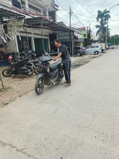 The image captures an outdoor scene depicting a man working on a scooter on the side of a street, likely at a motorcycle repair shop.

In the foreground, a man stands next to a black scooter (moped). He is a middle-aged individual with dark, short hair and appears focused on the scooter's handlebars and exposed front components. He is wearing a dark, short-sleeved t-shirt, dark track pants with white stripes on the sides, and light-colored flip-flops. The scooter is partially disassembled, with its front fairing removed, revealing wires and other internal parts. Its front wheel is turned slightly to the left.

Behind the man and to his left, there is an area that appears to be a motorcycle repair or service shop. Several other motorcycles, some partially disassembled, are visible on the dirt and gravel ground. Various motorcycle parts, including shiny exhaust pipes, are hanging from a metal rack under a makeshift awning that extends from the main building. A stack of tires and a blue plastic stool can also be seen in this area.

The main building on the left is a multi-story structure with a white facade and a brown roof line. Its ground floor features closed doors, possibly the entrance to the shop. Utility poles with numerous overhead wires crisscross the sky above the buildings and street.

The street itself is made of light-colored concrete or asphalt, showing some wear and tear, with small cracks and debris, including what appear to be cigarette butts, scattered on its surface. The road extends into the distance on the right side of the frame. Further down the street, various buildings, including one with a green facade, line the road. Several cars are parked along the curb, including a light blue minivan and a darker-colored car. Palm trees and other greenery are visible in the background against an overcast sky.

The overall impression is that of a bustling, everyday street scene in a town or city, with a focus on local commerce and vehicle maintenance. The sky is cloudy, suggesting a daytime setting without direct sunlight. The man's attire and the general setting hint at a location in a tropical or subtropical region, possibly Southeast Asia.