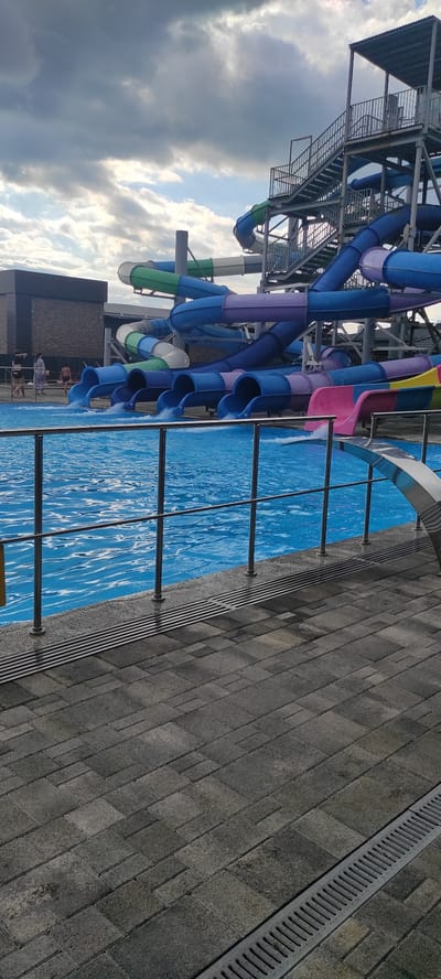 The image shows a water park with multiple slides and a swimming pool. The slides are brightly colored in blue, purple, green, and pink, and they descend from a tall metal structure. Several people are visible in the background, near the entrance of some slides or walking by the pool. The swimming pool is filled with clear blue water and is surrounded by a paved area. The sky is cloudy, with patches of blue sky visible. The overall scene depicts a recreational area on an overcast day. This location is in the city of Tulskiy, Russia.