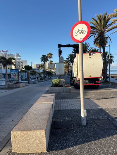 The image depicts a street scene in Adeje, Spain, with a clear blue sky and bright sunlight, suggesting it's daytime. The orientation of the image is portrait, with the ground occupying the bottom half and the sky the top. Palm trees are a prominent feature, lining the pedestrian walkway.
In the foreground, a round traffic sign with a red border and a white center indicates a prohibition for bicycles, with a pictogram of a bicycle crossed out. A lamppost supports the sign. To the right, a long, grey granite bench is situated on the pedestrian path, which is paved with grey tiles. Further down the path, a large digital billboard advertises "FUN SHOPPING" with images of various products.
A white truck with a large, light-colored cargo container is parked on the road next to the sidewalk. The truck has the company logo "PALFINGER" visible on its side. Traffic lights are visible in the mid-ground, suggesting this is an intersection or a controlled area. In the background, several multi-story white buildings are visible, some with balconies. The sea can be seen in the distance at the top of the image. The overall atmosphere is bright and sunny, indicative of a pleasant day in a coastal town. No people are clearly visible in the frame.