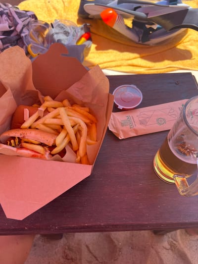 An overhead shot depicts a meal of a hot dog and french fries in a brown paper box, placed on a dark wooden table. To the right of the box is a clear glass mug filled with a light-colored beverage, likely beer, with a foamy head. A small plastic container with red sauce sits to the left of the box. A paper wrapper, with "TESSERA" and "EAT DIFFERENT" printed on it, lies between the sauce container and the beer mug. In the background, a yellow towel and some beach accessories are visible. The scene is set on a sandy surface, suggesting an outdoor location such as a beachside eatery in Νέα Φλογητά, Greece.