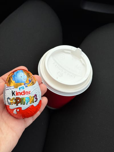 A hand holds a Kinder Surprise egg wrapped in foil. The foil is mostly red and silver, with the Kinder logo clearly visible in white and a cartoon character in blue and yellow on one side. The egg is partially obscuring a white paper lid of a red coffee cup. The lid has some text and markings on it, including what appears to be "IPA X UNIT". The background is dark, suggesting an indoor setting or possibly a car interior, with a textured black surface. The lighting is diffused, but the overall scene suggests a casual, everyday moment. The text "Усть-Сыны, Russia" indicates the general location.