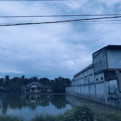The image captures a quiet, overcast landscape in what appears to be Payameuneng, Indonesia, featuring a still body of water, buildings, and dense tropical foliage. The upper third of the frame is dominated by a light blue-grey sky with soft, diffused clouds, suggesting an overcast day or perhaps early morning/late afternoon. Several dark, horizontal power lines traverse the sky, slightly sagging.
A tranquil expanse of water occupies the mid-ground, serving as a mirror to the sky and the structures along its banks. On the right, a large, grey, industrial-looking concrete building with a flat roof stretches along the water's edge. Its long side features multiple regularly spaced, rectangular openings or vents. The base of the wall appears to be constructed from a rougher material, possibly stone or textured concrete, and a section with dark railings is visible towards the right end of the building. The structure's reflection is clearly cast upon the calm water.
To the left, across the water and nestled among lush greenery, stands a residential house. It features a distinctive, possibly corrugated roof and a small porch. The house itself appears to be a light, faded color, perhaps cream or pale yellow, with subtle decorative details on its facade. Dense tropical vegetation, including numerous palm trees, forms a verdant backdrop to the house and extends throughout the background, characteristic of the region. The immediate foreground shows blurry green foliage, possibly indicating the photograph was taken from a moving perspective.
No people are visible in the scene, and there is no discernible text. The overall atmosphere is one of stillness and the blending of human construction with the abundant natural environment under a muted sky.