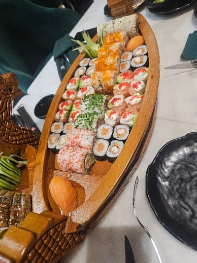 A variety of sushi rolls fill a wooden boat-shaped platter. The rolls feature different fillings and toppings like avocado, salmon, and colorful roe. Cucumber slices garnish one section. A black plate sits above the platter, with a fork and knife nearby. The setting suggests a restaurant with green seating and a marble-like table. Some of the boat's exterior is decorated with carved patterns.