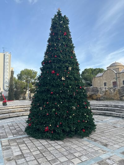 The image, captured in Agios Dometios, Cyprus, features a very tall, conical Christmas tree dominating the foreground of an outdoor public space. The artificial tree is lush with dark green foliage and richly decorated with an abundance of red and green spherical ornaments, glittering gold star-shaped decorations, and gold bow-shaped accents. Small white lights are visible strung across its branches, and a prominent golden star crowns the tree.

To the right of the tree, an old, historic-looking stone building with a distinct dome and small windows is partially visible, appearing to be situated on a raised, rocky foundation with stone steps leading up to it. Further back and to the left, parts of a contemporary building with a flat roof and an antenna can be seen, along with additional green trees. Near the modern building, a piece of red and black machinery, possibly a lift or crane, is partially visible.

The ground around the base of the Christmas tree is paved with square, patterned tiles in shades of grey and beige, interspersed with a section of lighter blue-grey tiles forming a border. The scene is illuminated by natural daylight under a clear to partly cloudy blue sky, indicating fair weather. No people are present in the image, but the elaborately decorated tree signals the Christmas season or a festive public event. The overall impression is one of serene holiday preparation. There is no visible text.