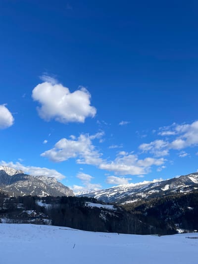 A vibrant blue sky filled with scattered cumulus clouds dominates the scene. Below, snow-covered mountains rise sharply, dotted with trees and glimpses of structures. Patches of white snow extend into the foreground. The image is oriented sideways, revealing a clear contrast between the bright sky and the rugged mountainous landscape.