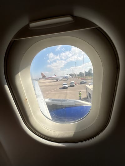 The image is an interior view from an airplane window, looking out onto an airport tarmac. In the foreground, a portion of the airplane's wing, painted blue and white, is visible. Beyond the wing, on the tarmac, a commercial airplane with "Arkia" branding is parked. Several vehicles, including vans and cars, are also present on the tarmac. Two individuals wearing high-visibility vests are standing near the parked aircraft and a service vehicle. The sky is blue with scattered clouds. The scene appears to be at Ben Gurion Airport, located in Yehud Monosson, Israel, given the presence of Arkia aircraft and the general airport infrastructure.