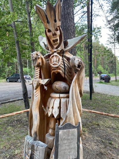 A weathered, carved wooden sculpture resembling a skeletal figure in a crown and armor stands in a wooded area. The sculpture is detailed, with visible ribs, a skull-like face, and a sword held in its left hand. A wooden plaque is attached to the base of the sculpture. In the background, there is a dirt path, several cars parked, and a few people walking. The environment appears to be a park or outdoor museum, likely the Tal'tsy Architectural-Ethnographic Museum, a historical settlement located near Irkutsk, Russia, known for its preserved wooden structures and crafts. The lighting suggests it is daytime, possibly late afternoon, with a soft, diffused light filtering through the trees.