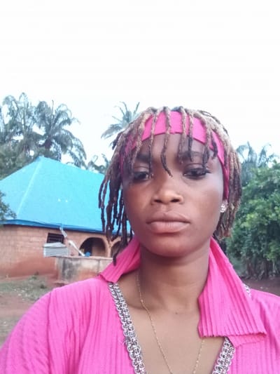 The image is a portrait of a woman, oriented vertically but rotated 90 degrees clockwise. A dark-skinned woman is prominently featured in the foreground, looking directly at the camera with a neutral, calm expression. She has full lips and her eyes are accentuated with dark eyeliner or mascara. Her hair is styled in dreadlocks, some of which appear to have pink highlights or are covered by a matching pink headwrap. She is wearing a bright, pleated pink top or dress with a silver-colored, decorative trim along the neckline. A thin, gold-colored chain necklace is visible around her neck.
The background depicts an outdoor setting during daytime, under what appears to be an overcast sky. Behind the woman, a building with a light blue roof and terracotta-colored walls is visible, along with some corrugated metal or scaffolding nearby. To her left and right, lush green trees and palm fronds are present, indicating a tropical or subtropical environment. The overall scene suggests a residential or semi-rural area in Ogbe, Nigeria, as provided by the context. No visible text is present in the image.