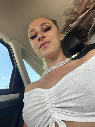 The media file is an upside-down selfie taken inside a car. The subject is a young woman with glitter eyeshadow and false eyelashes, wearing a white textured dress and a pearl necklace. She is looking at the camera with a slight smile. The car's interior is visible, including the roof lining and a portion of the passenger side window. The background shows a bright blue sky, suggesting it is daytime with clear weather. There are no discernible location cues in the image other than the specified location context of Prijeradi, Montenegro, and the presence of a VW sticker on the window.